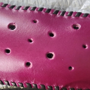 May include: A close-up of a burgundy leather strap with a black stitched border and multiple holes. The strap has a silver metal buckle on one end.