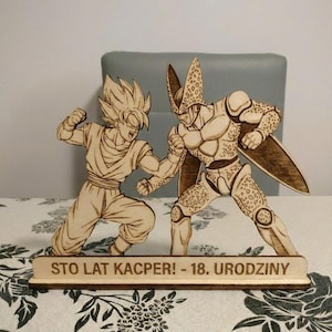 May include: Wooden laser-cut figures of two Dragon Ball Z characters in a fighting pose. The figures are light brown with detailed etching. A wooden base below the figures reads "STO LAT KACPER! - 18. URODZINY".