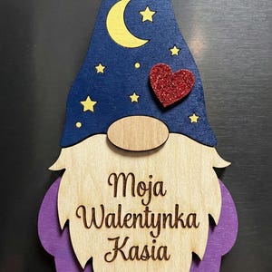 May include: Wooden gnome decoration with a blue hat featuring a yellow crescent moon and stars. A red glitter heart is above the wooden beard. The text "Moja Walentynka Kasia" is engraved on the beard.
