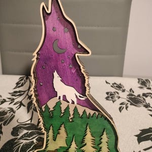 Puede incluir: Obra de arte de madera en capas que representa la silueta de un lobo aullando contra un cielo nocturno púrpura con una luna creciente y estrellas. El lobo está sobre una colina sobre un bosque de árboles verdes y beige. La obra es de madera en capas.