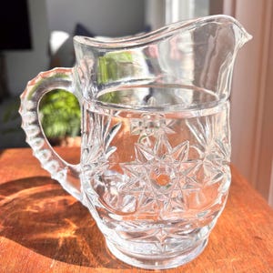 May include: A clear glass pitcher with a starburst pattern and a handle. The pitcher has a spout for easy pouring and is likely used for serving beverages. The design is etched into the glass, adding a decorative element to the functional item.