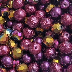 May include: Close-up of a collection of beads in various shapes and colors. The beads are primarily shades of purple and gold, with some iridescent and faceted designs. The beads are round and have holes for stringing.