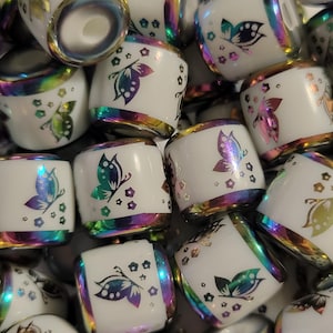 May include: A collection of white ceramic beads with a cylindrical shape. Each bead features a butterfly design in iridescent colors and is accented with a gold rim. The beads are scattered, creating a visually appealing arrangement.