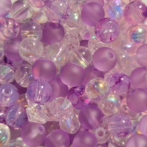 May include: A collection of purple and clear beads. The beads vary in size and shape, including round, faceted, and irregular forms. Some beads have a matte finish, while others are transparent or iridescent, creating a mix of textures and light reflections.