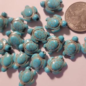 May include: A collection of turquoise turtle-shaped beads with a quarter for size comparison. The beads have a textured surface and are a light blue color with brown accents. These beads are ideal for jewelry making.