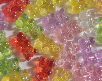 May include: A collection of colorful, translucent bear-shaped beads in various shades of red, orange, yellow, green, pink, and purple. These small, decorative beads are scattered across a white surface.