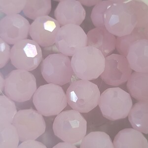 Pu&ograve; includere: Primo piano di un gruppo di perline di vetro sfaccettate rosa pallido. Le perline hanno un leggero bagliore iridescente.