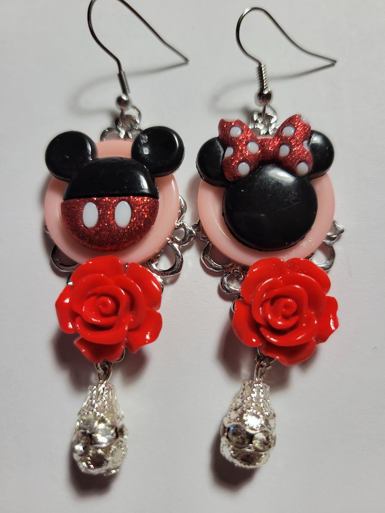 May include: A pair of dangle earrings featuring a red and black Mickey Mouse and Minnie Mouse design with a red rose and silver charm on each earring.