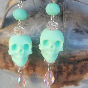 May include: A pair of mint green skull earrings with silver-tone hooks. Each earring features a skull charm, faceted beads, and a teardrop-shaped pink crystal. The earrings are designed for pierced ears.