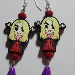May include: A pair of dangle earrings featuring a cartoon character design. Each earring has a silver hook, a red bead, a character charm with blonde hair and a red and purple outfit, and a purple teardrop bead.