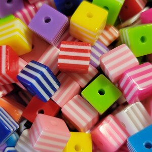 May include: A collection of colorful, striped, square-shaped beads with a hole in the center. The beads are made of plastic and have a glossy finish.