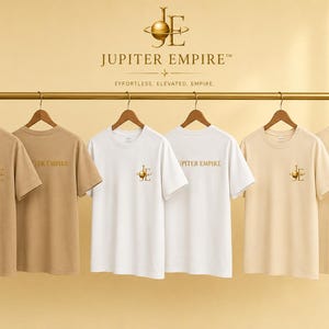 Könnte beinhalten: Sechs kurzärmelige T-Shirts in Beige-, Weiß- und Cremetönen hängen an einer goldenen Kleiderstange. Die Shirts tragen den Schriftzug JUPITER EMPIRE und ein goldenes Planetenlogo. Einige zeigen das Logo auf der Brust, andere den Markennamen.