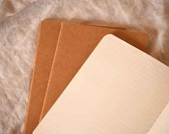 May include: A brown notebook with lined pages is open and lying on top of two other brown notebooks. The notebooks are on a light brown fabric surface.