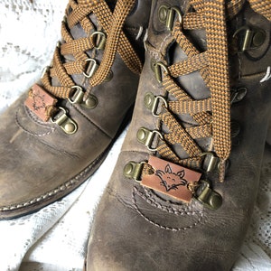 Könnte beinhalten: Ein Paar braune Lederstiefel mit braunen und orangefarbenen Schnürsenkeln. Die Stiefel haben ein Fuchskopfdesign auf den Lederschnürsenkeln.