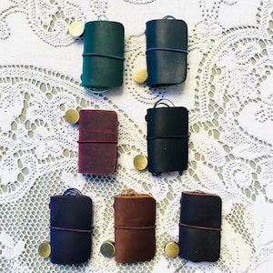 Puede incluir: Seis pequeños cuadernos encuadernados en cuero de varios colores, incluyendo verde, azul, marrón y burdeos. Cada cuaderno tiene un cierre de metal dorado y está atado con una cuerda.