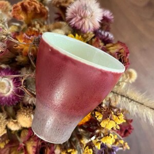 May include: A handmade ceramic mug with a gradient of colors, transitioning from a deep red at the top to a light gray at the base. The interior is a pale green. The mug is set against a backdrop of dried flowers.