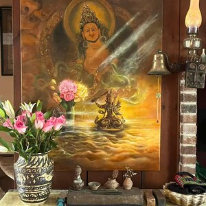 May include: An ornate painting of a deity with a golden halo, set against a warm, golden background. A bronze statue and a vase of pink roses and white lilies sit below. A decorative bell and a lamp are mounted on the wooden wall.