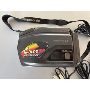 Op de afbeelding: Een donkergrijze Panasonic Palmcorder IQ camcorder met een zwarte draagriem. Het apparaat is voorzien van een regenboogkleurige Color Viewfinder-sticker, x10 full auto operation-tekst en een VHS-C PV-IQ403-label. Inclusief een zwarte stroomkabel.