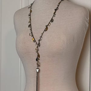 May include: A silver-toned necklace with a Y-shaped design. The necklace features small, irregularly shaped charms in gold, silver, and clear tones. A tassel of silver chains hangs from the bottom. The necklace is displayed on a mannequin.