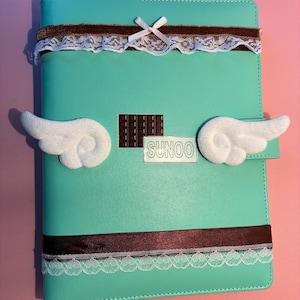Op de afbeelding: Een mintgroen notitieboek met een bruin lint en witte kanten rand aan de boven- en onderkant. De kaft is versierd met twee witte pluche vleugels, een kleine bruine chocoladereepbedel en een rechthoekig label met de tekst SUNOO in het zwart.
