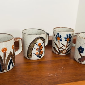 May include: Four ceramic mugs with speckled white exteriors and brown handles. Each mug features a unique floral design in shades of orange, blue, and brown. The mugs are arranged on a wooden tray.