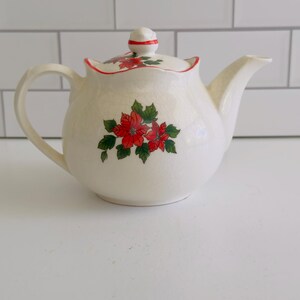 May include: A cream-colored ceramic teapot featuring a delicate red poinsettia floral pattern with green leaves on the body and lid. The teapot has a curved handle, a tapered spout, and a lid with a red-trimmed edge and a small knob handle.