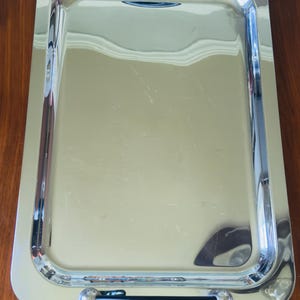 May include: A rectangular, silver-colored serving tray with rounded corners and a raised edge. The tray has two handles with black grips and decorative silver accents. The tray is sitting on a brown wooden surface.