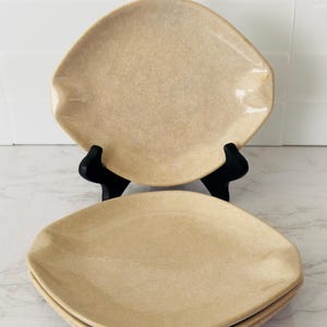 May include: A stack of four beige, diamond-shaped plates. The plates have a speckled texture and are displayed on a white surface. One plate is propped up on a black stand, showcasing the unique shape and design.