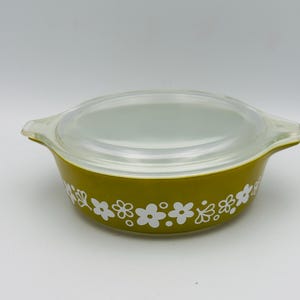May include: An olive green Pyrex casserole dish with a clear lid. The dish features a white floral pattern around the exterior. The handles are integrated into the design, and the lid fits snugly on top. A vintage kitchenware item.