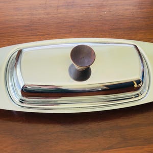 May include: A rectangular, silver-colored butter dish with a rounded, off-white border. The lid features a small, round, dark brown wooden knob. The dish is set on a dark brown wooden surface.