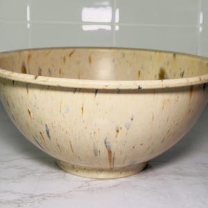 May include: A large, speckled mixing bowl with a cream-colored base and a wide rim. The bowl features a speckled pattern with brown, blue, and gray accents. The bowl is made of ceramic and is suitable for kitchen use.