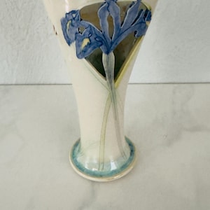 May include: A cream-colored ceramic vase with a flared rim and a circular base. The exterior features a raised, hand-painted blue iris flower design with green stems extending toward the base. The rim and base are accented with a light blue glaze.