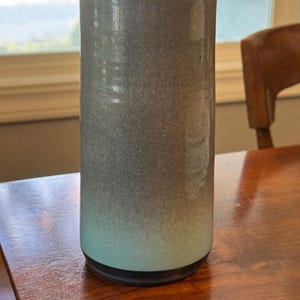 May include: A tall, cylindrical ceramic vase with a wavy top. The vase has a gradient color scheme, transitioning from a dark gray at the top to a light teal at the base. The vase is approximately 10 inches tall.