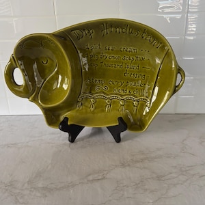 May include: Olive green ceramic serving dish in the shape of an elephant. The dish has the words "Dip Hindustani" and a recipe for the dip. The dish is on a black stand.