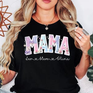May include: Black t-shirt with the word "MAMA" in a patchwork design with floral accents. Below the word "MAMA" are the names "Leo", "Steve", and "Olivia". The shirt is a crew neck style.