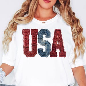 May include: White t-shirt with the word "USA" in red and blue denim-style letters. The letters have a stitched border, giving a vintage, patriotic look. The shirt is a casual style.