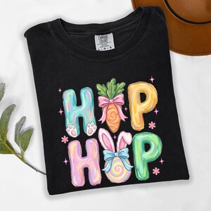 Puede incluir: Camiseta negra con las palabras "HIP HOP" en letras de burbujas coloridas. Las "H" tienen patas de conejo, las "O" son zanahorias y un huevo de Pascua, y las "P" son amarillas y verdes. La camiseta es de la marca Comfort Colors.
