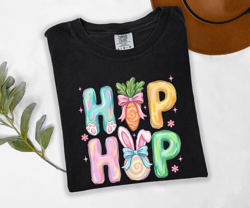 Puede incluir: Camiseta negra con las palabras "HIP HOP" en letras de burbujas coloridas. Las "H" tienen patas de conejo, las "O" son zanahorias y un huevo de Pascua, y las "P" son amarillas y verdes. La camiseta es de la marca Comfort Colors.