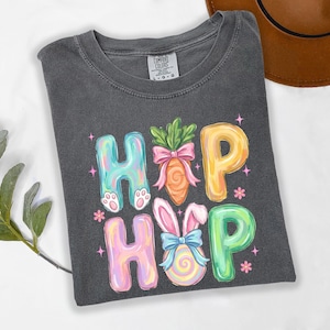 Lifestyle flat lay product image of a soft comfort t-shirt featuring colorful pastel “Hip Hop” lettering decorated with Easter-themed illustrations. The design includes bunny ears, carrot graphic, ribbon bows, small flowers, and playful sparkles. The shirt is neatly folded on a minimal neutral background with subtle decorative props, creating a bright, cheerful spring aesthetic ideal for Easter apparel and seasonal Etsy product listings.