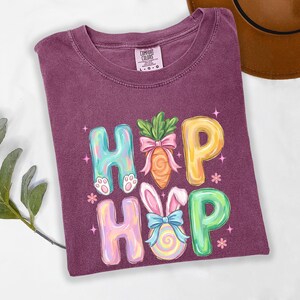 Puede incluir: Una camiseta morada con las palabras "HIP HOP" en letras de burbujas coloridas. Las "H" tienen patas de conejo, las "O" orejas de conejo y las "P" est&aacute;n decoradas con una zanahoria y un huevo de Pascua. La camiseta es de la marca Comfort Colors.