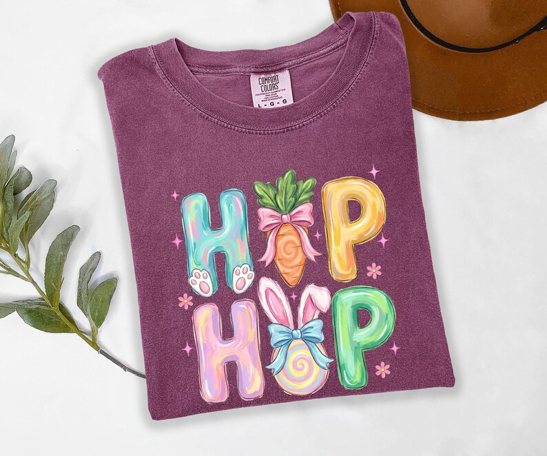 Puede incluir: Una camiseta morada con las palabras "HIP HOP" en letras de burbujas coloridas. Las "H" tienen patas de conejo, las "O" orejas de conejo y las "P" est&aacute;n decoradas con una zanahoria y un huevo de Pascua. La camiseta es de la marca Comfort Colors.