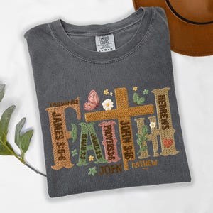 May include: Dark gray t-shirt with the word "FAITH" embroidered in a textured, colorful design. The design includes a cross, butterfly, and floral accents, with Bible verses incorporated into the letters.