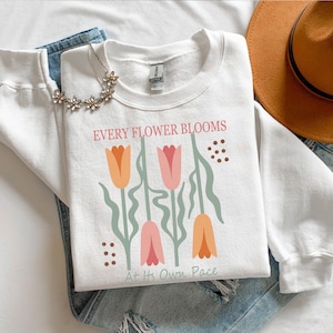 May include: White sweatshirt with the text "EVERY FLOWER BLOOMS At Its Own Pace" and a floral design in orange, pink, and green. The sweatshirt is paired with blue jeans, a necklace, and a brown hat.