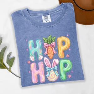 Puede incluir: Una camiseta azul Comfort Colors con un gr&aacute;fico colorido "HOP HOP". El dise&ntilde;o presenta huellas de conejito, una zanahoria con un lazo rosa y un huevo de Pascua con orejas de conejo y un lazo azul. Las letras son de colores pastel.