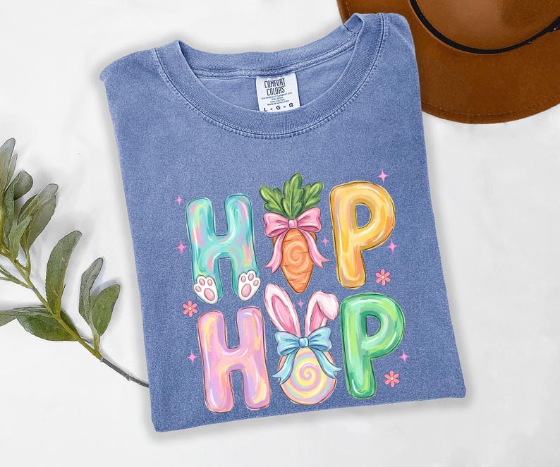 Puede incluir: Una camiseta azul Comfort Colors con un gr&aacute;fico colorido "HOP HOP". El dise&ntilde;o presenta huellas de conejito, una zanahoria con un lazo rosa y un huevo de Pascua con orejas de conejo y un lazo azul. Las letras son de colores pastel.