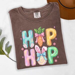 Puede incluir: Camiseta marr&oacute;n con un gr&aacute;fico colorido "Hopp". El dise&ntilde;o presenta letras pastel, una zanahoria con un lazo rosa y un conejito con un huevo. La camiseta es de la marca Comfort Colors.