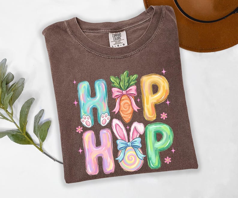 Puede incluir: Camiseta marr&oacute;n con un gr&aacute;fico colorido "Hopp". El dise&ntilde;o presenta letras pastel, una zanahoria con un lazo rosa y un conejito con un huevo. La camiseta es de la marca Comfort Colors.