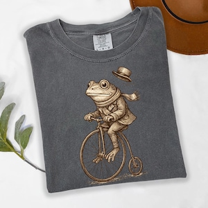 May include: Dark gray t-shirt featuring a whimsical illustration of a frog riding a penny-farthing bicycle. The frog is dressed in a coat, scarf, and hat. The shirt is a Comfort Colors brand.
