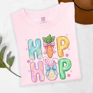 Puede incluir: Una camiseta rosa claro Comfort Colors con la palabra "HIP HOP" en letras coloridas y caprichosas. Las "H" tienen patas de conejo, las "O" son una zanahoria y un huevo de Pascua, y las "P" son amarillas y verdes. La camiseta est&aacute; decorada con estrellas y flores.