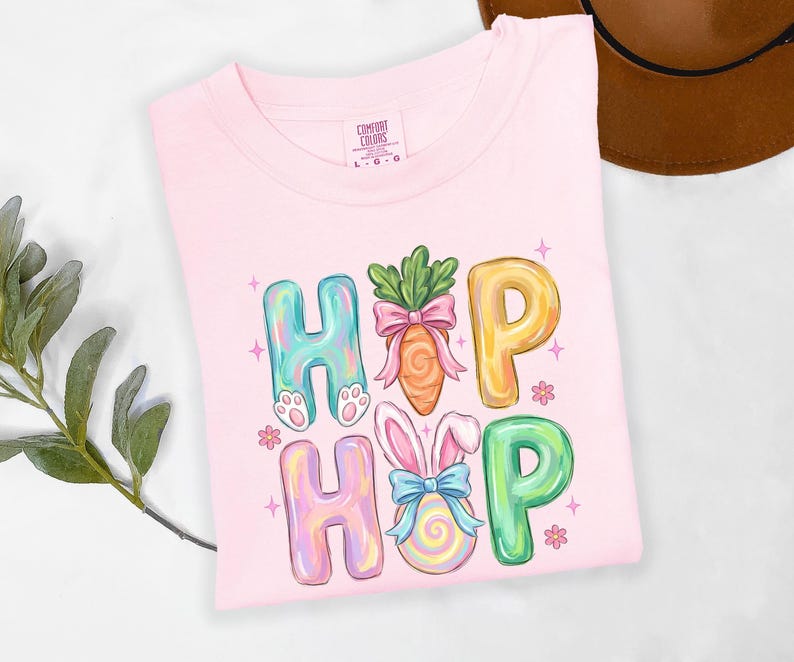 Puede incluir: Una camiseta rosa claro Comfort Colors con la palabra "HIP HOP" en letras coloridas y caprichosas. Las "H" tienen patas de conejo, las "O" son una zanahoria y un huevo de Pascua, y las "P" son amarillas y verdes. La camiseta est&aacute; decorada con estrellas y flores.
